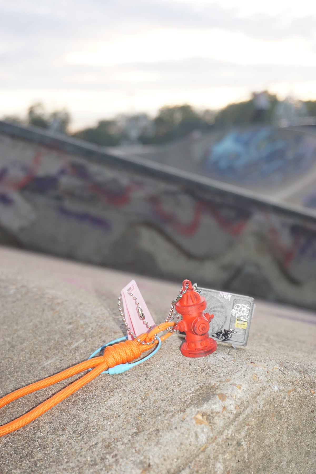 A small red fire hydrant keychain with an orange cord and a pink tag lies on a concrete ledge in front of a blurred skatepark ramp at sunset.