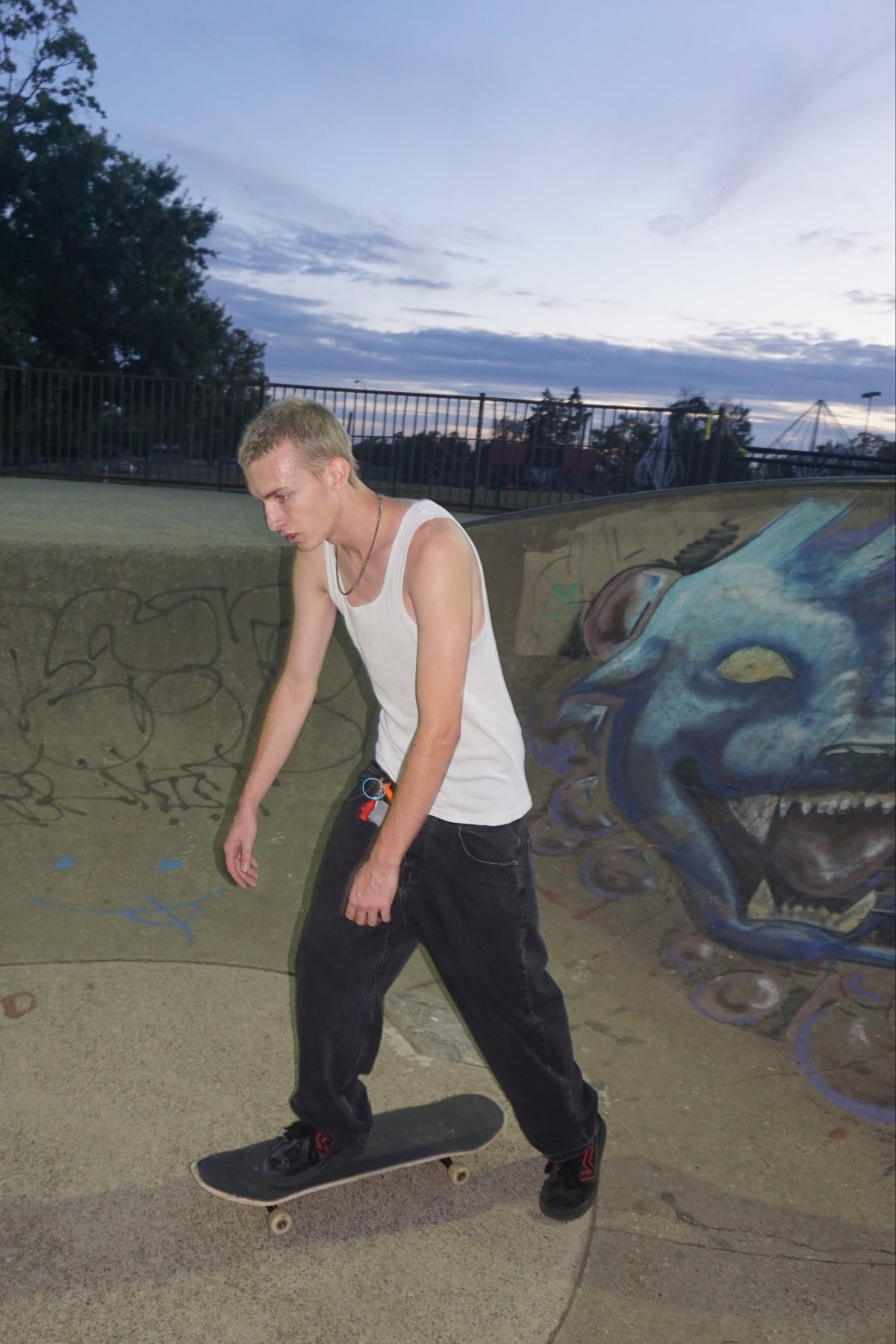A man in a white tank top and dark pants balances on a skateboard inside a concrete skatepark bowl with graffiti murals under a twilight sky.